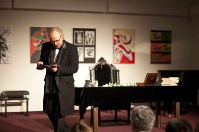 Der Schauspieler im langen schwarzen Gehrock liest auf der Bühne ein Blatt, hinter ihm der Tisch mit Schachfiguren und Büchern