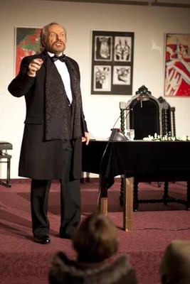 Der Schauspieler im Gehrock deklamiert mit erhobener Hand vor dem Bühnentisch und einem geschnitzten Lehnstuhl