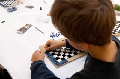 Kind legt in der Museumswerkstatt schwarze und weiße Mosaiksteine zu einem Schachbrett auf einer Holzplatte, daneben blaue Steinchen