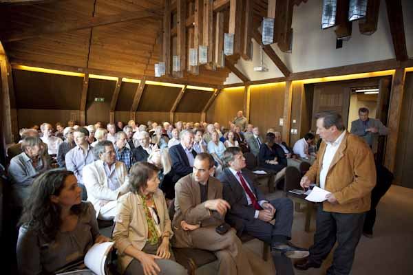 Voll besetzter Saal mit Holzdecke zur Ausstellungseröffnung in Grafing, vorn steht ein Mann mit Manuskript vor dem Publikum