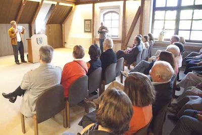 Ein Redner spricht neben dem Pult mit Stadtwappen zu den Gästen, die in Stuhlreihen unter Dachschrägen sitzen