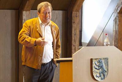 Ein Redner in senfgelber Wildlederjacke spricht neben einem Rednerpult mit Wappen, das einen Löwen zeigt