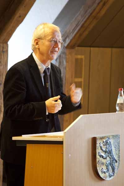 Ein Redner mit Brille im dunklen Anzug spricht gestikulierend am Pult mit Stadtwappen