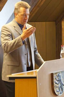Ein Redner im hellgrauen Anzug spricht am Rednerpult mit dem Löwenwappen, daneben eine Wasserflasche
