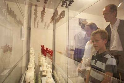 Ein Junge und Erwachsene betrachten eine Vitrine mit Elfenbein-Schachfiguren in Weiß und Rot