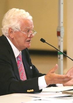Ein weißhaariger Referent mit Brille und rosa Krawatte spricht gestikulierend ins Tischmikrofon