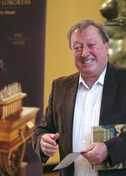 Ein lächelnder Mann im braunen Sakko hält einen Ausstellungskatalog, im Hintergrund ein Schachexponat und ein Banner