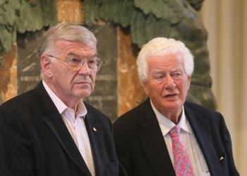 Zwei ältere Herren in dunklen Sakkos sitzen nebeneinander und hören zu, dahinter ein Steindenkmal mit Inschrift