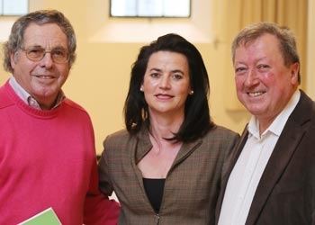 Ein Mann im roten Pullover, eine Frau im karierten Blazer und ein Mann im Sakko lächeln gemeinsam in die Kamera