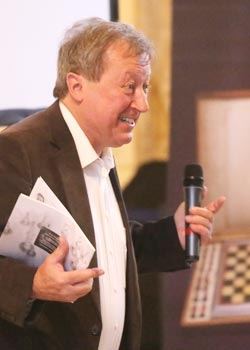 Ein Redner mit Handmikrofon und Papieren unterm Arm spricht, im Hintergrund unscharf ein Schachbrett