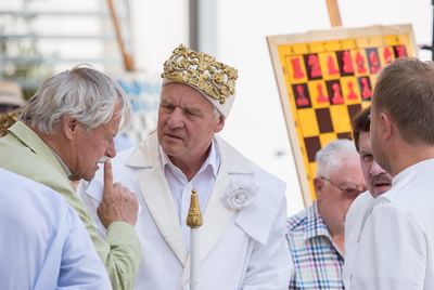 Weißer König mit goldener Krone und Zepter im Gespräch mit einem Großmeister, hinten das große Demonstrationsbrett