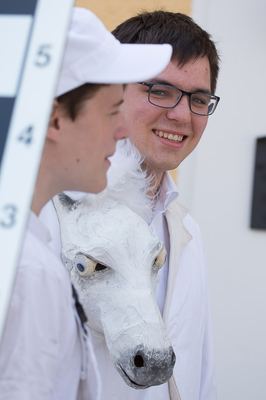 Junger Mann lächelnd neben einem Mitspieler, der einen weißen Pferdekopf aus Pappmaché als Springer-Kostüm trägt
