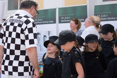 Kinder in schwarzen Latzhosen und Kappen hören einem Mann im Schachbretttrikot zu, dahinter Plakate „50 Jahre Schachunion“