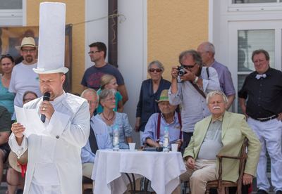 Grußwort des weißen Turms vor dem Kommentatorentisch mit weißer Decke, dahinter Gäste und ein Fotograf