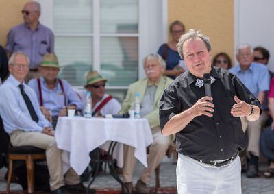 Redner im schwarzen Hemd mit Fliege spricht ins Mikrofon, hinter ihm Ehrengäste am weiß gedeckten Kommentatorentisch