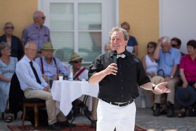 Redner mit Mikrofon wendet sich lächelnd ans Publikum, das an der gelben Hausfassade sitzt