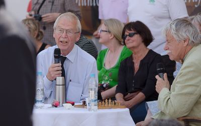 Ein Großmeister kommentiert mit Mikrofon am weiß gedeckten Tisch mit Analysebrett, neben ihm sein Kommentatorkollege