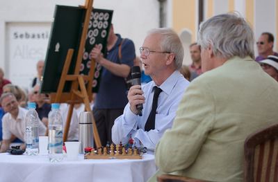 Großmeister erläutert am Kommentatorentisch einen Zug, im Hintergrund ein Helfer am Demonstrationsbrett