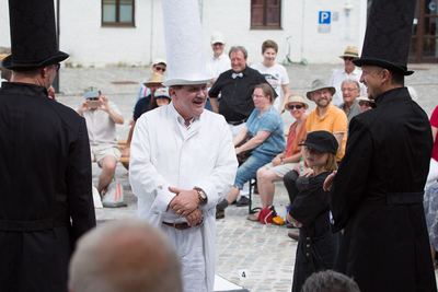 Weißer Turm mit hohem Hut lacht zwischen zwei schwarzen Läufern, im Hintergrund Zuschauer auf dem gepflasterten Hof