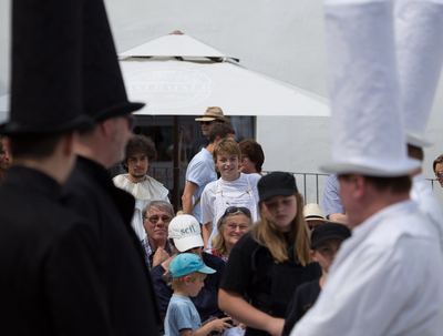 Schwarze und weiße Figuren mit hohen Hüten rahmen den Blick auf Zuschauer unter einem Sonnenschirm