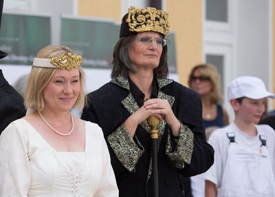 Weiße Dame mit Perlenkette und schwarzer König mit Goldkrone und Zepter nebeneinander, rechts ein weißer Bauer