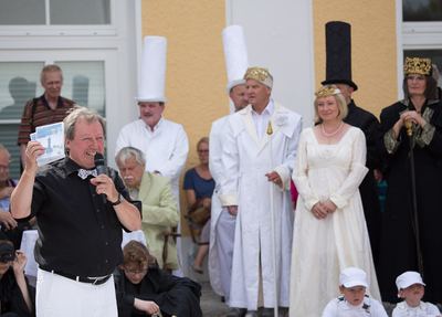 Moderator hält eine Karte hoch, hinter ihm reihen sich Türme, Königspaare und ein Läufer beider Farben auf