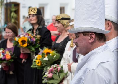 Weißer Turm mit hohem Hut im Profil, daneben Königinnen und König mit Sonnenblumensträußen