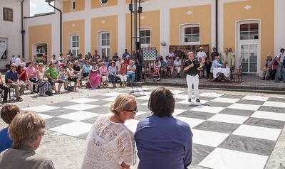 Überblick über den Theaterhof: großes schwarz-weißes Spielfeld, Moderator mit Mikrofon und Publikum vor gelber Fassade