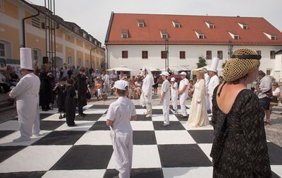 Weite Sicht über das Schachbrett im Hof: weiße Bauern, weiße Dame und schwarze Figuren vor dem Gebäude mit rotem Dach