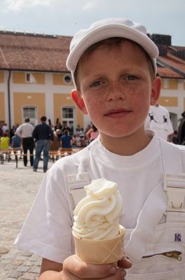 Junge im weißen Bauernkostüm mit Kappe hält eine Waffel mit Softeis, hinten der sonnige Hof