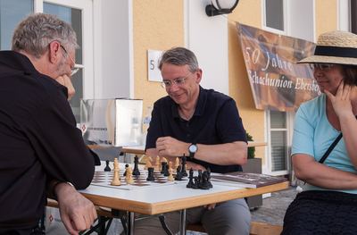 Nach der Aufführung: zwei Männer spielen an einem Tisch Schach, eine Zuschauerin mit Strohhut schaut zu