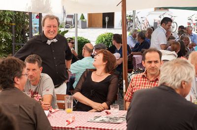 Der Moderator mit Fliege steht lachend zwischen den Gästen an den karierten Tischen im Biergarten
