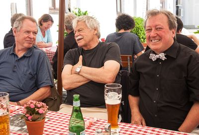Ein Großmeister sitzt lachend mit verschränkten Armen zwischen Gästen am Biergartentisch