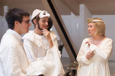 Weiße Dame mit goldverziertem Stirnband im Gespräch mit zwei weißen Bauern mit Halskrause und Maske vor dem Auftritt