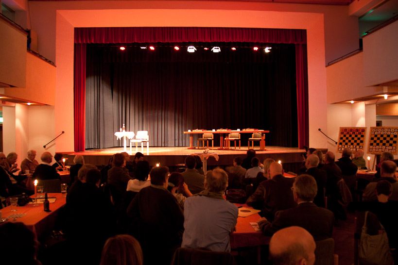 Blick über das Publikum an Kerzentischen auf die noch leere Bühne der Stadthalle Grafing mit Tischen, Schachbrettern und Demobrettern