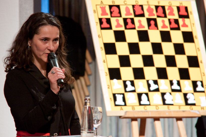 Die Moderatorin spricht ins Mikrofon, hinter ihr ein großes Demonstrationsschachbrett in Grundstellung auf einer Staffelei