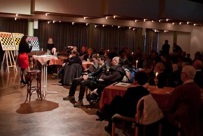 Gut besetzter Saal mit Kerzen auf den Tischen; links spricht die Moderatorin vor drei Demobrettern zum Publikum