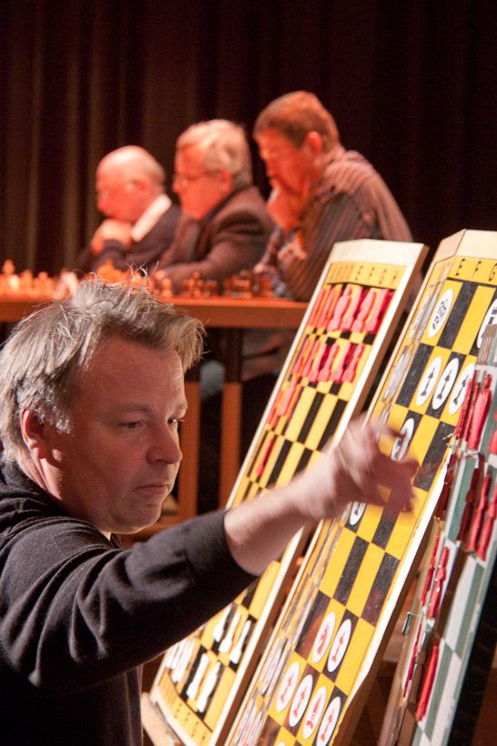 Ein Großmeister setzt eine Figur am gelb-schwarzen Demobrett, hinter ihm grübeln die drei Herausforderer an ihren Partien