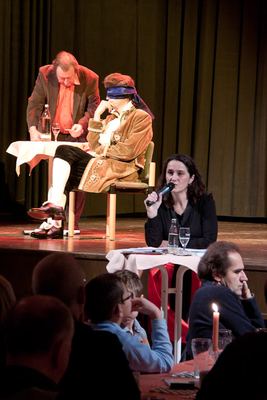 Die Moderatorin spricht am Stehtisch ins Mikrofon, hinter ihr auf der Bühne notiert der Sekundant neben dem blinden Darsteller