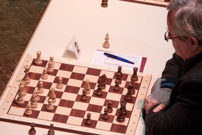 Stellung an Brett 2 im Mittelspiel, ein Herausforderer mit verschränkten Armen betrachtet das Brett, vor ihm das Partieformular