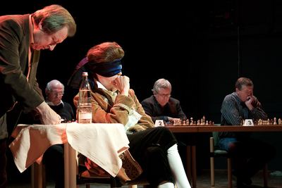 Der Sekundant notiert, während der Darsteller mit Augenbinde die Hand ans Kinn legt; hinten die Herausforderer an Brett 2 und 3