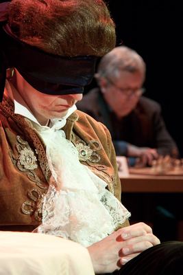 Der blinde Darsteller mit gefalteten Händen, bestickter Samtjacke und Spitzenjabot, hinten unscharf ein Herausforderer