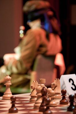 Weiße und schwarze Figuren an Brett 3 im Fokus, im unscharfen Hintergrund der kostümierte Darsteller mit Augenbinde