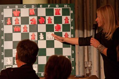 Die FIDE-Meisterin zeigt mit Mikrofon in der Hand auf eine Stellung am grün-weißen Demobrett mit roten und weißen Figuren