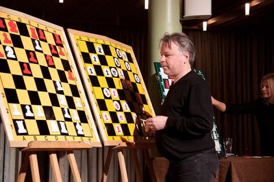 Der Großmeister hält Figuren in der Hand und tritt vom gelb-schwarzen Demobrett zurück, rechts setzt die FIDE-Meisterin einen Zug