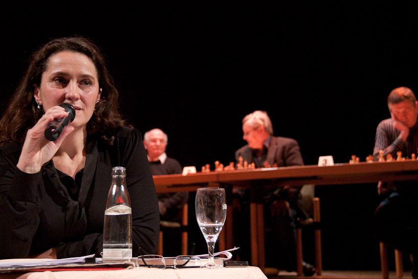Die Biografin spricht am Tisch ins Mikrofon, hinter ihr auf der Bühne die drei Herausforderer an ihren Brettern