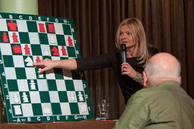 Die FIDE-Meisterin zeigt mit ausgestrecktem Arm auf eine weiße Figur am grün-weißen Demobrett, vorn ein Zuschauer