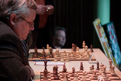 Ein Herausforderer mit Brille blickt auf sein Brett, hinter der Spieltafel steht der Sekundant, rechts die Demobretter