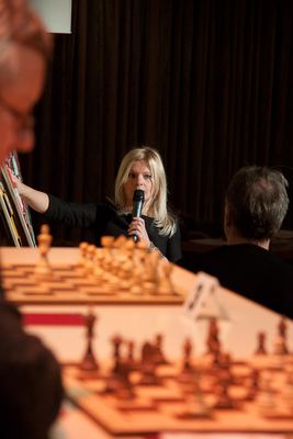 Die FIDE-Meisterin spricht ins Mikrofon und zeigt zum Demobrett, vorn unscharf die Figuren auf der Spieltafel