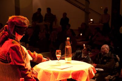 Der Darsteller mit Augenbinde sitzt im warmen Bühnenlicht am Tisch mit Wasserflasche und Glas, dahinter das Publikum
